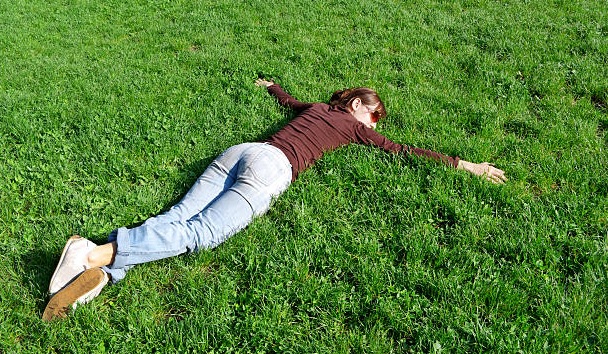 Devojka u patikama, farmerkama i braon majici lezi u travi rasirenih ruku licem okrenutim ka zemlji