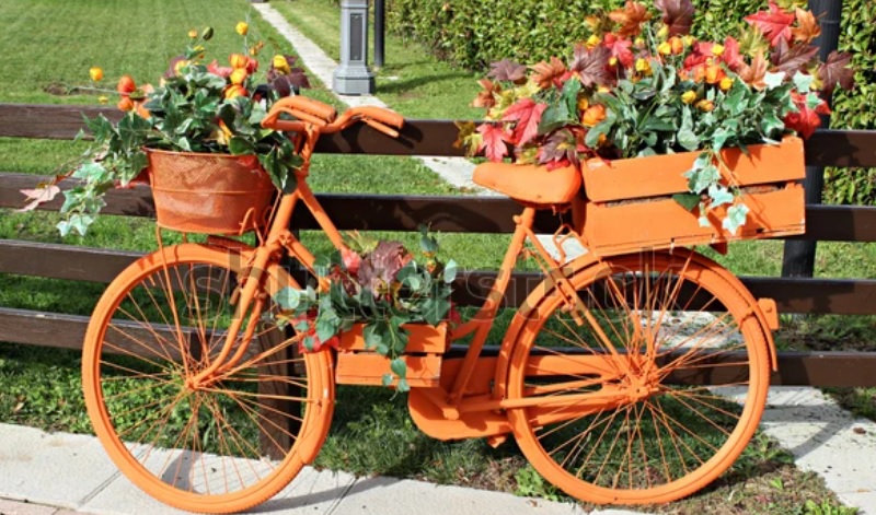 Na travniku uz drvenu ogradu stoji narandžasti bicikl sa dve gajbice i korpom punim cvecem
