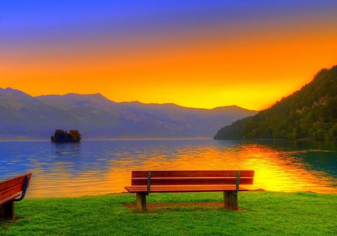 Drvena klupa na zelenoj travi uz obalu mirnog jezera sa pogledpm na planine i odsjaj sunca