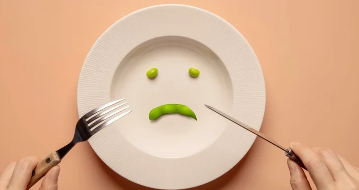 Viljuska i noz naslonjeni na krem tanjir u kome su dva zrna graska kao oci i neoljusten grasak kao usta